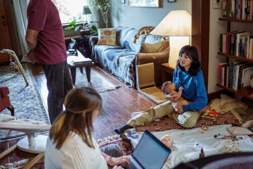 In a living room, an early interventionist sits on the floor with a laptop, taking notes during a home visit, while a mother holds her baby on a floor cushion nearby. Another baby lies on a blanket in the foreground surrounded by toys, and a man stands in the background near a baby gym toy. Bookshelves, a couch, and a lamp fill out the lived-in space.