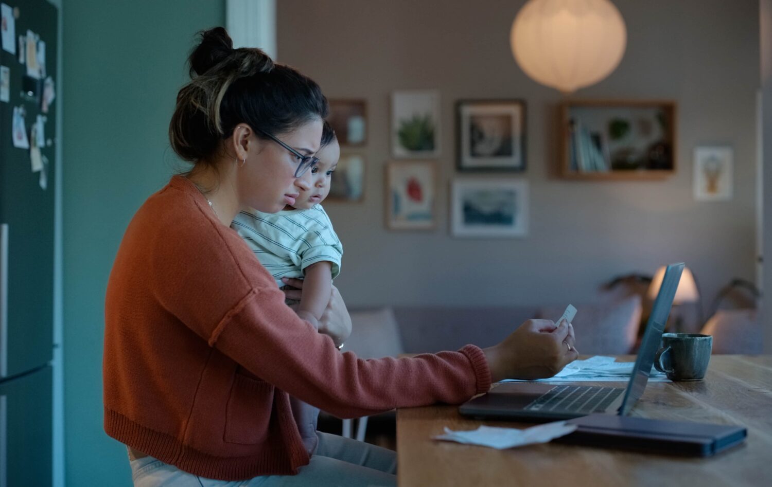 A woman in a rust-colored sweater holds a small child while looking at her laptop at a wooden dining table, surrounded by warm lighting and framed pictures.