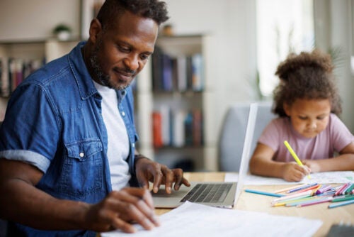 An adult Black male uses a laptop while a young child sits at the table next to him coloring.