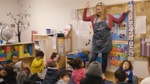 A teacher stands in front of young children sitting on a rub. The teacher is raising her right hand and pointing at a picture schedule.