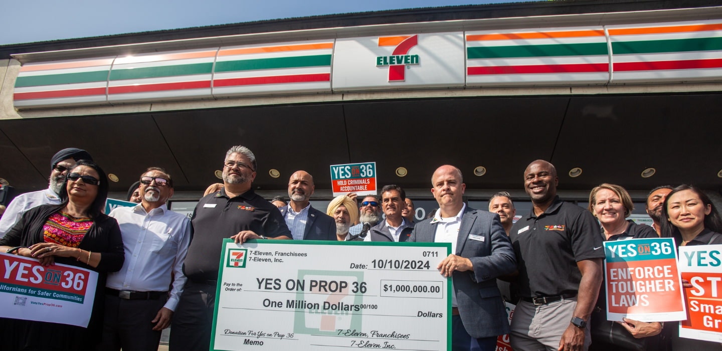 A group of diverse individuals celebrates a one million dollar donation for Proposition 36 in front of a 7-Eleven store, holding signs that read 