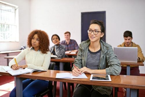 College students sit at desks in a classroom, looking toward the front of the room and taking notes in notebooks and on a laptop.