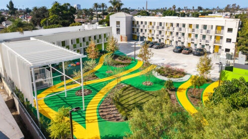 Housing development showcasing a U-shaped apartment complex with four floors and a courtyard with bright green and yellow pavement, and trees scattered throughout.