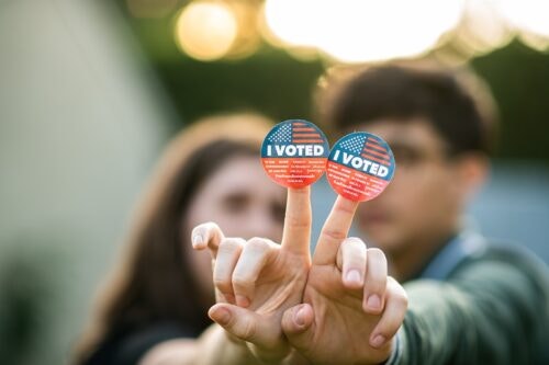 Portrait of two women holding out their hands together with the I voted stickers attached to their fingers for the viewer to see. Their fingers form the “v” for victory and peace sign.