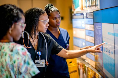 Three women in health care attire are engaged with an interactive display in a health care setting, one pointing at the screen while two others observe.