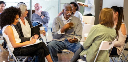 A diverse group of six people are seated in a circle, engaged in a discussion, with an older man in a gray cardigan speaking animatedly at the center.