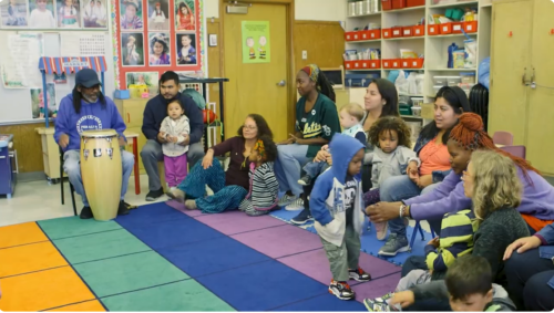 Young children and their caregivers sit in a semicircle in a preschool classroom while a man plays a drum.