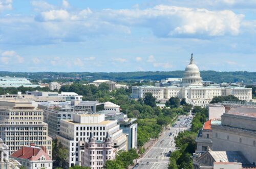 Panoramic view of the United States Capitol surrounded by modern buildings and tree-lined streets, under a blue sky with clouds.