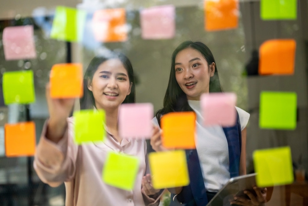 Two women stand in front of a wall with Post-its covering it, participating in a brainstorm.