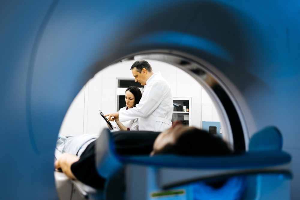A radiological technologist shows computed tomography scan results to an apprentice while a patient lies down in the machine.