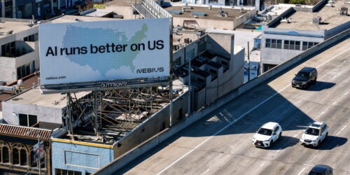 Rooftop billboard reading “AI runs better on us” above an elevated roadway with several cars.