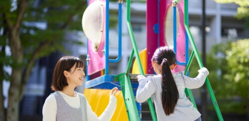 A woman stands beside a colorful playground smiling as a child climbs a green ladder.