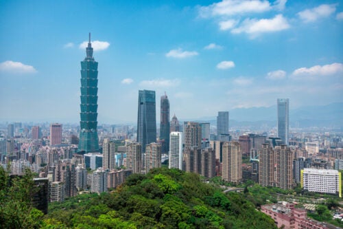 The skyline of Taipei, Taiwan, with green foliage in the foreground, skyscrapers in the middle, and mountains in the distant background