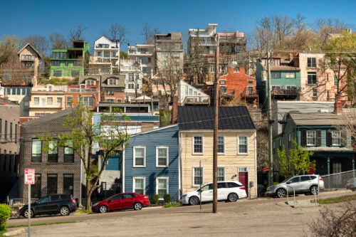 In the foreground are a line of parked cars along a hilly street. In the background on the hillside are several rows of historic attached houses