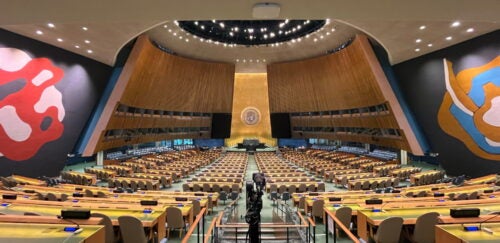 Empty international assembly hall with rows of desks and seats facing a central podium, wood-paneled walls, colorful abstract murals, and a camera in the center aisle.