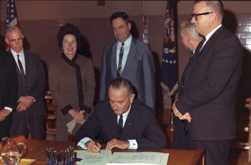President Johnson signs bill with three people standing over him