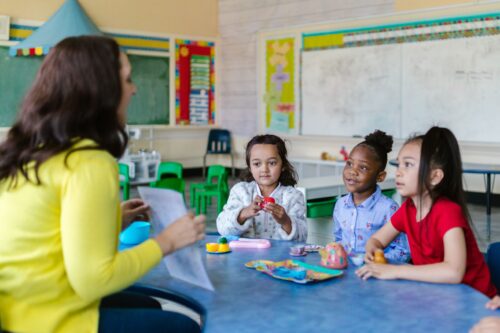 Three young children sit at a table in a classroom looking at their teacher, who is holding up a piece of paper.