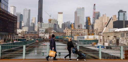 A man and a woman pushing a stroller walk along a canal path in an urban area, with modern skyscrapers and construction sites visible in the background.