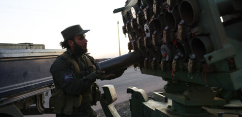 A person in military uniform loads a rocket into a multiple rocket launcher system at dusk, with an armored vehicle visible in the background.