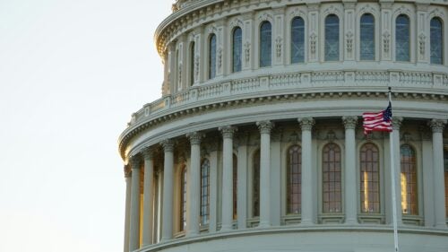 Close-up image of the Capitol Building dome in fading light, with the US flag waving in the forefront.