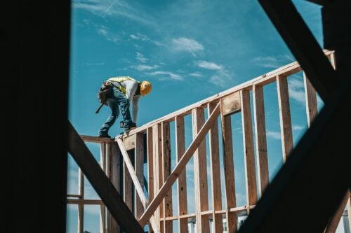 Construction worker standing on top of a wall frame of a building under construction, with blue sky in the background.