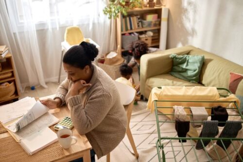 Black mother sitting at table, looking at finances while children play in the background.