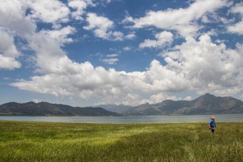 Person with a colorful backpack stands in a grassy field beside a lake, facing rugged mountains beneath a cloud-filled blue sky.