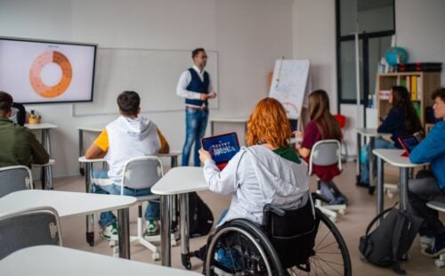 A classroom with multiple students with a focus of a girl with red hair in a wheel chair looking at an ipad