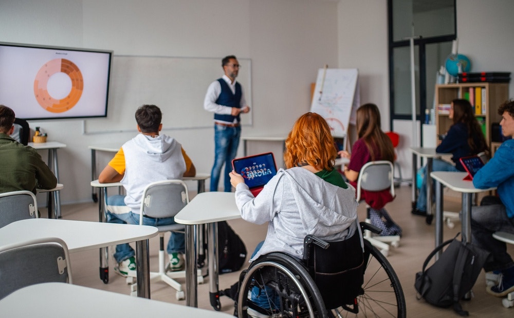 A classroom with multiple students with a focus of a girl with red hair in a wheel chair looking at an ipad