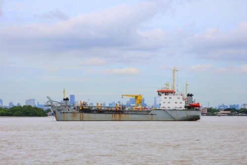 Large gray cargo ship with yellow deck cranes traveling along a river, with a city skyline in the background.