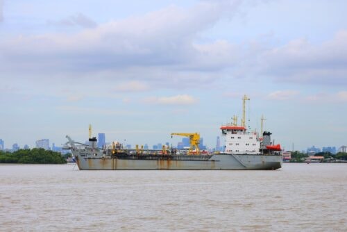 Large gray cargo ship with yellow deck cranes traveling along a river, with a city skyline in the background.