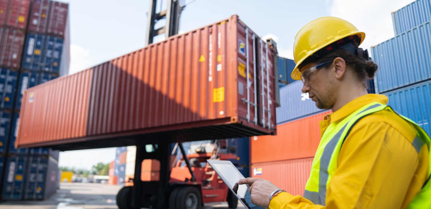 Worker in safety gear uses a tablet while a container handler lifts a red shipping container in a shipping yard.