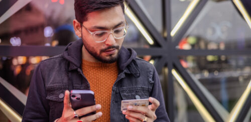 Man looking at a smartphone while holding a payment card in a brightly lit, geometric indoor setting.