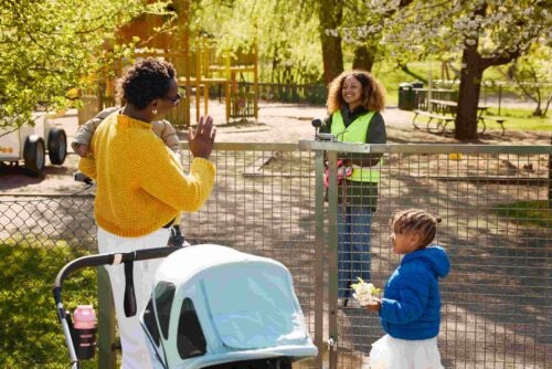 People interact across a fenced playground, with a child holding white flowers beside a stroller.