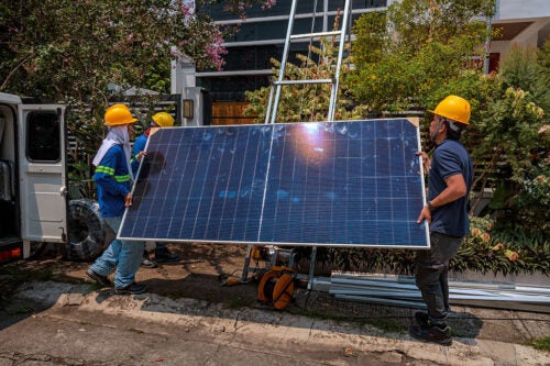 Three workers in yellow hard hats are lifting a solar panel in front of a tree-lined house
