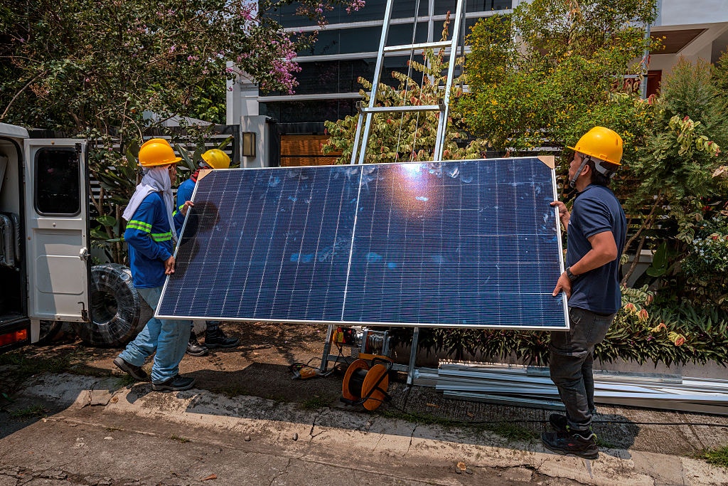 Three workers in yellow hard hats are lifting a solar panel in front of a tree-lined house