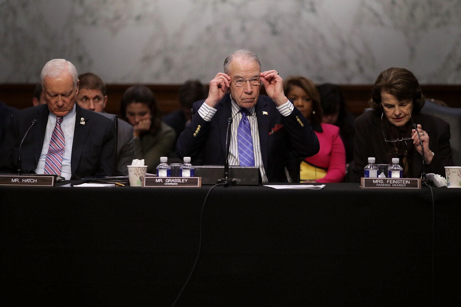 Two older men and an older woman are seated in a row before a table containing nameplates, water bottles and microphones. The center man and the woman are both putting on glasses.