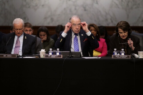 Two older men and an older woman are seated in a row before a table containing nameplates, water bottles and microphones. The center man and the woman are both putting on glasses.