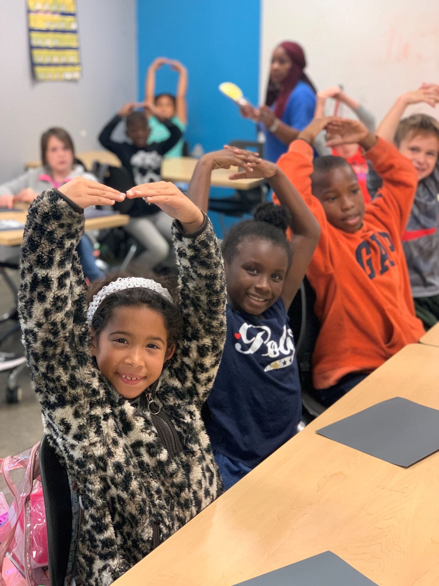 Children in an afterschool classroom put their hands in an O-shape over their heads and smile at the camera.