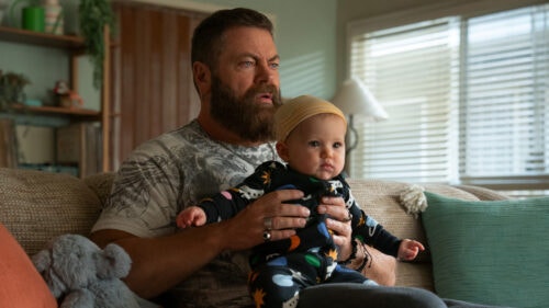 Bearded man holding a baby on a couch in a sunlit living room.