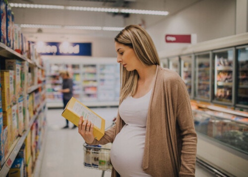 Pregnant woman holding a yellow packaged item in a brightly lit grocery-store aisle.