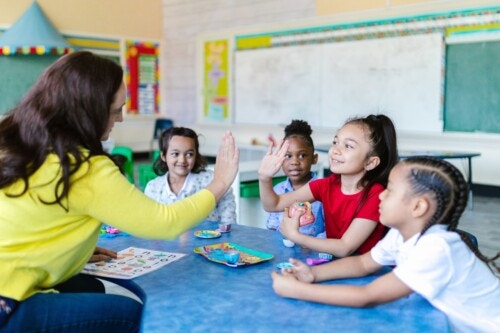 A teacher in a yellow sweater high-fives a smiling young girl at a classroom table, while three other children look on. Toy tea cups, plates, and a letter-matching game are spread across the table.