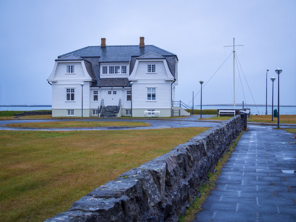 A large white house with a gray roof sits on the waterfront. In front is a wet slate path and a lawn of brown grass. The sky is clouded over.
