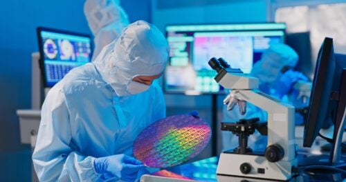 Asian male technician in sterile coverall holds wafer that reflects many different colors with gloves and check it at semiconductor manufacturing plant