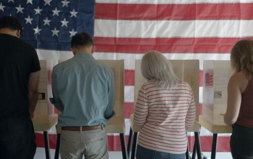 The backs of voters as they cast their ballots in front of an American flag.