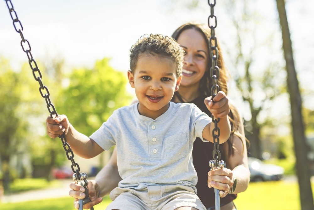 A long haired woman pushes her young son in a light colored t-shirt on a swing set. Both are smiling.
