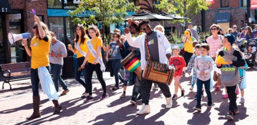 A diverse group of people marching in a vibrant street parade, led by a person with a megaphone and another playing a drum.